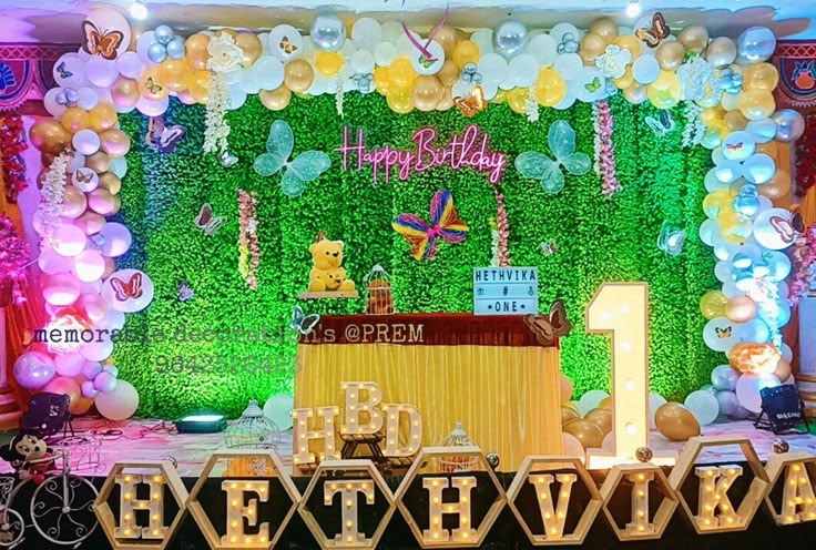 Butterfly theme first birthday decoration in Hyderabad with a green grass wall, pastel balloon arch and light-up name letters