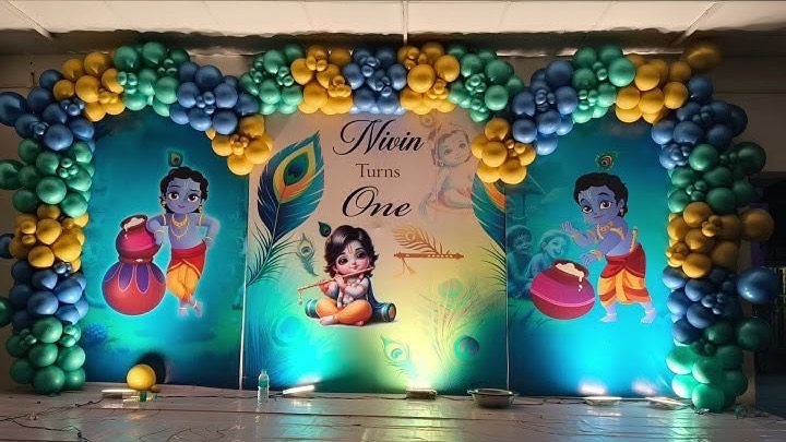 Krishna theme first birthday decoration in Hyderabad with a peacock feather backdrop and blue, green and gold balloon arch